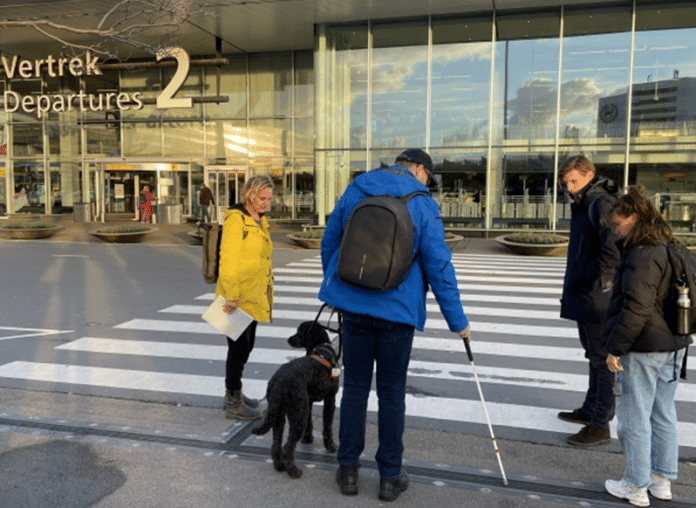 Frouck de Boer en Peter Waalboer aan het werk om de toegankelijkheid van Schiphol te verbeteren voor mensen met een visuele handicap. Ze staan buiten voor Vertrekhal 2 en bespreken een geleidelijn bij een voetgangersoversteekplaats